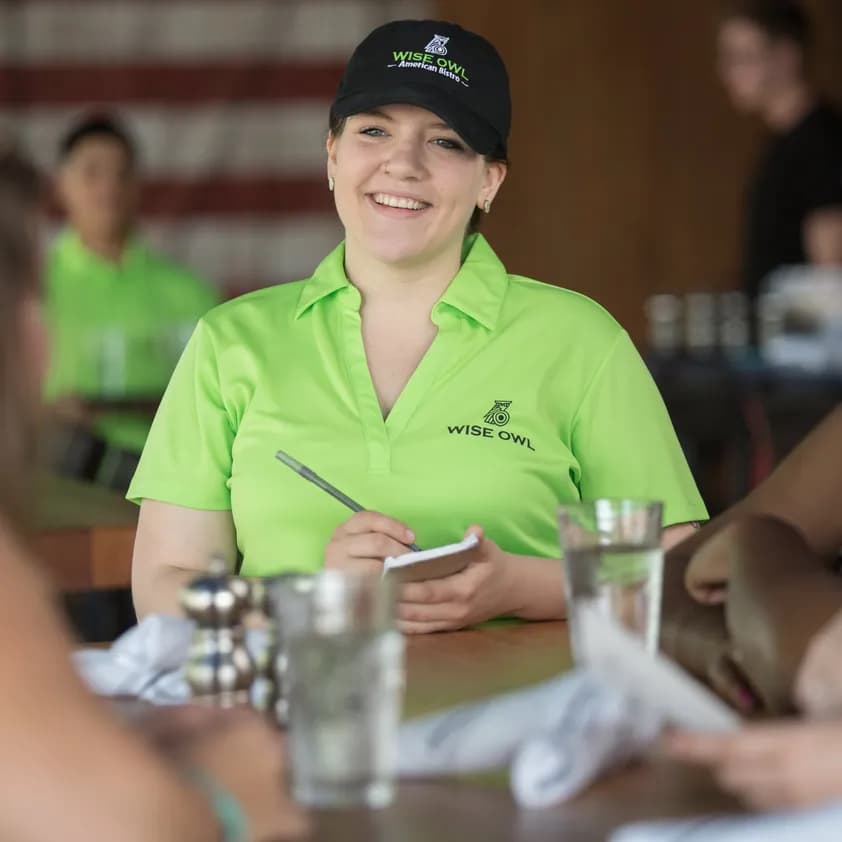 A server wearing branded hat and polo shirt, takes a customer order in the Wise Owl American Bistro.