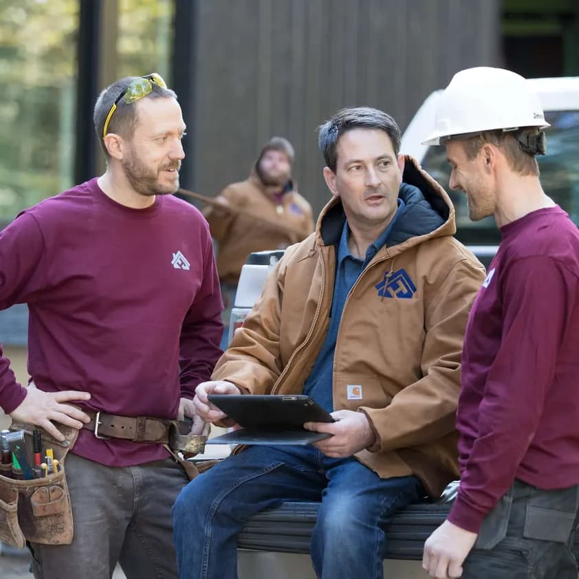 A group of construction workers wearing custom long sleeve t-shirts and jackets, sit on the back of a flatbed truck discussing work.