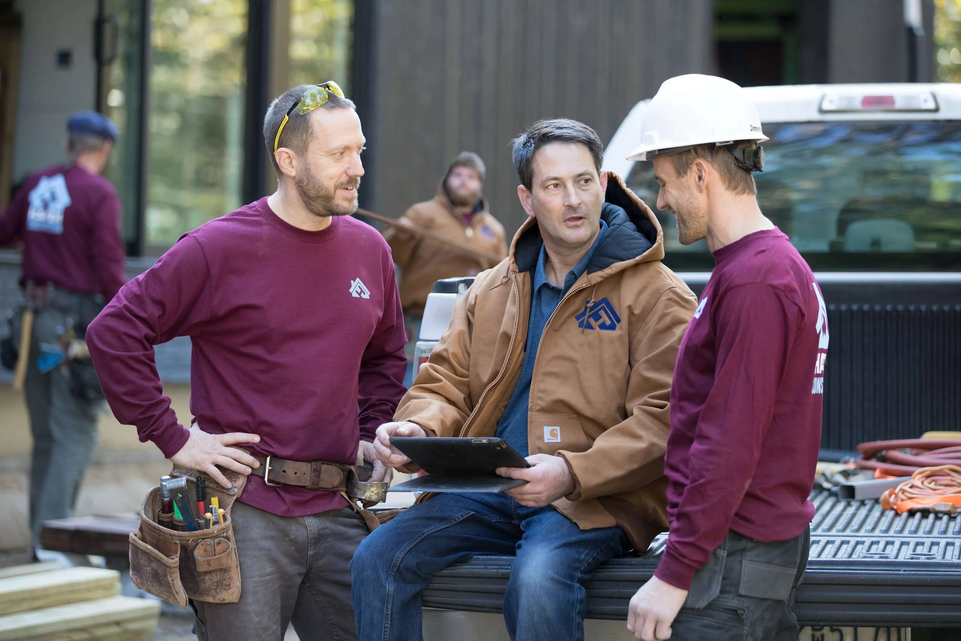 A group of construction workers wearing custom long sleeve t-shirts and jackets, sit on the back of a flatbed truck discussing work.