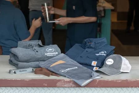 A table features folded branded uniforms for a brewery including polo shirts, button up shirts, waist apron, trucker hat, and bottle opener.