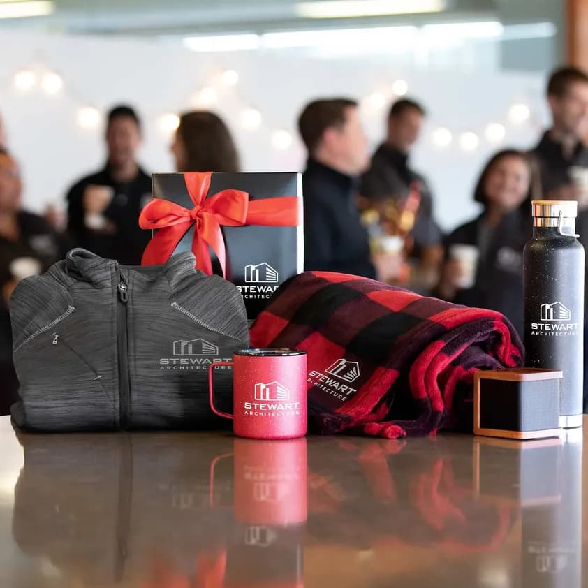 Branded company gifts including custom logo jacket, mug, blanket, and bottle displayed on a table with coworkers in the background.