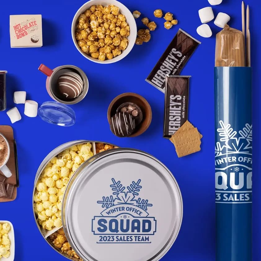 A laydown of custom holiday themed treats are scattered on the table, including a tin of popcorn, hot chocolate and smores kit.